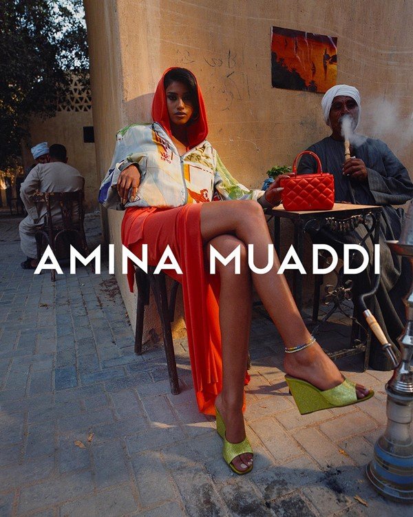 Campanha de divulgação da marca de sapatos Amina Muaddi. Na foto, é possível ver uma mulher sentada em uma mesa em uma área externa com um homem mais velho fumando narguilé. Ela usa uma sandália de salto verde, um vestido laranja e uma camisa branca de seda por cima