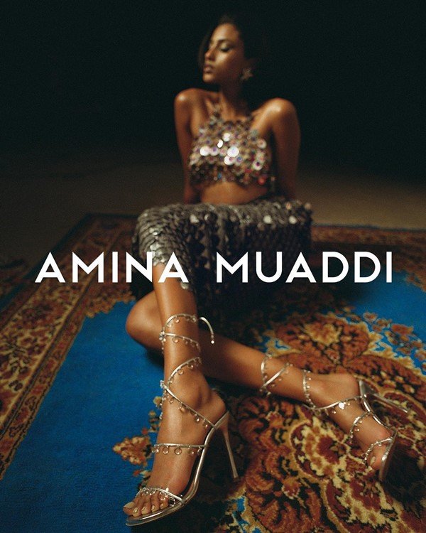 Campanha de divulgação da marca de sapatos Amina Muaddi. Na foto, é possível ver uma mulher jovem e morena, árabe, posando em um tapete étnico. Ela usa um conjunto de top e calça de brilho prata e uma sandália, também de prata, com tiras até o meio da perna e pedras preciosas aplicadas à corda
