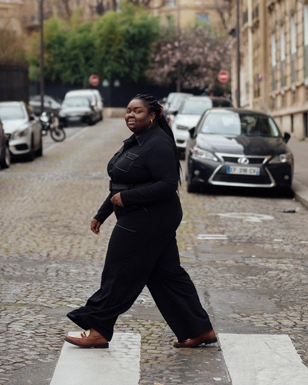 Mulher jovem e negra, de cabelos longos trançados, posando para foto em uma faixa de pedestre nas ruas de Londres. Ela usa um macacão jeans preto e um sapato mocassim de couro marrom e branco