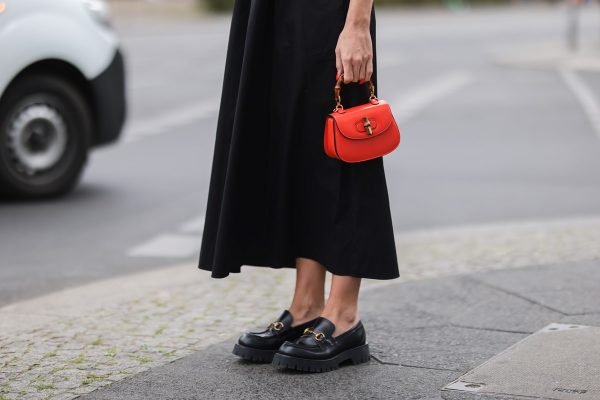 Mulher branca andando na rua de vestido longo preto, um sapato mocassim preto de couro e uma bolsa de mão laranja, da Gucci, com alça de bambu