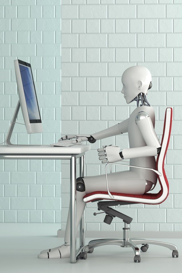 Color photo.  A robot sits and uses a computer
