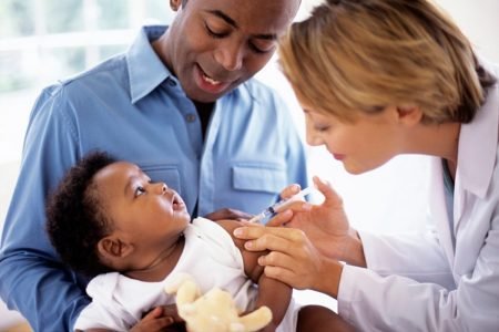 Foto colorida mostra pai segurando bebê enquanto mulher o vacina - Metrópoles