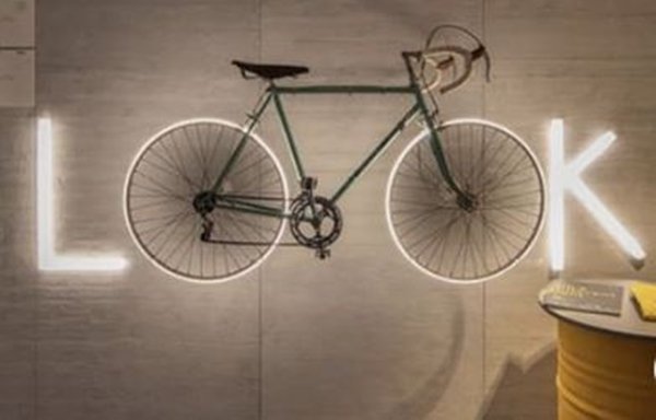 Foto colorida de uma parede escrita look em led. Uma bicicleta está sobre a palavra e as suas rodas compõem o nome