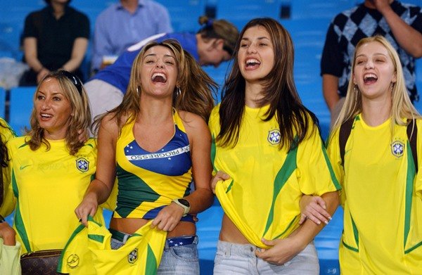 Quatro meninas jovens e brancas, de cabelo liso, torcendo pela seleção brasileira de futebol na Copa do Mundo de 2002. Todas usam a camisa verde e amarela 