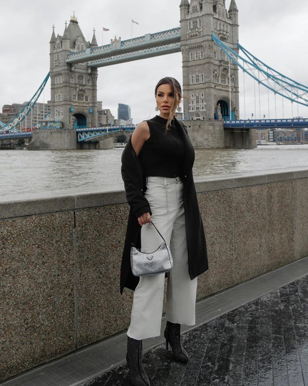 Mulher jovem e branca, com cabelo liso longo, posando para foto perto da London Bridge, na capital da Inglaterra. Ela usa uma regata preta de gola alta, uma calça jeans branca, um cardigan preto por cima e uma bolsa prata
