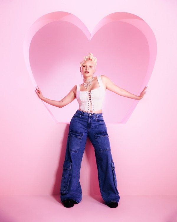 Campanha de divulgação da coleção de roupas da C&A em parceria com a cantora Duda Beat. Na foto, é possíverl ver Duda Beat, uma mulher branca, jovem e com cabelo loiro platinado em um fundo rosa. Ela veste um top corset branco e uma calça jeans