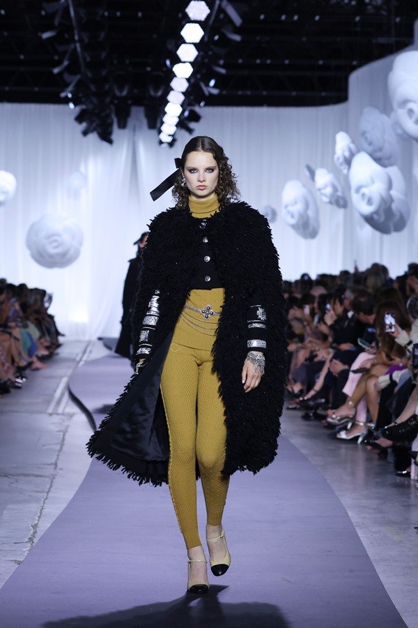 Modelo branca e jovem, de cabelo curto e cacheado, desfilando na passarela da marca francesa Chanel, durante a semana de moda. Ela usa um conjunto de casaco e calça legging, ambos amarelo mostarda, e por cima um sobretudo preto.