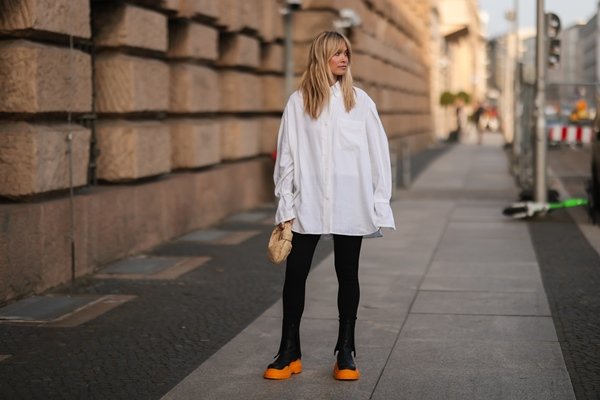 Mulher branca e jovem, de cabelo liso loiro, posando para foto nas ruas de Paris. Ela usa uma camiseta branca de botão, calça legging preta e uma bota coturno preta com o solado tratorado laranja