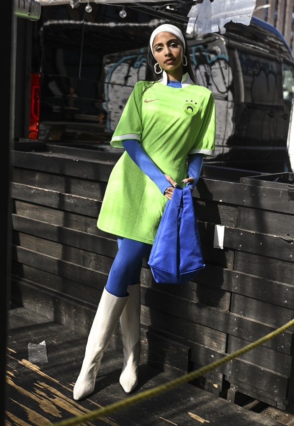 Mulher morena e jovem, de cabelo liso castanho, posando para foto nas ruas de Nova York. Ela usa uma calça legging e uma blusa de manga longa, ambas em um tom forte de azul, e por cima um vestido verde neon. 