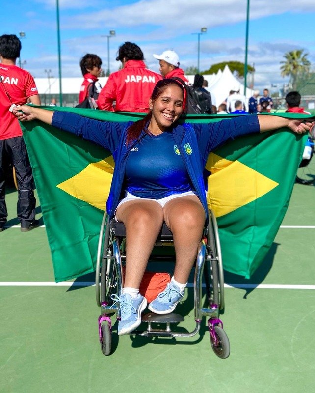 Jade em cadeira de rodas segurando bandeira do Brasil e sorrindo para foto