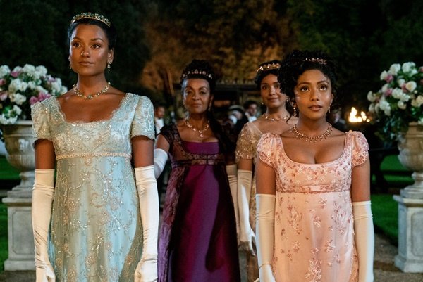 Cena da série Bridgerton, da Netflix, em que é possível ver quatro mulheres negras, de cabelo preso com coroas, e vestidos de época com renda e luvas