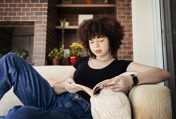 Mulher negra de pele clara, com cabelo cacheado curto, sentada em um sofá bege. Ela usa uma camiseta preta, calça jeans e um relógio smart. Ela está lendo um livro