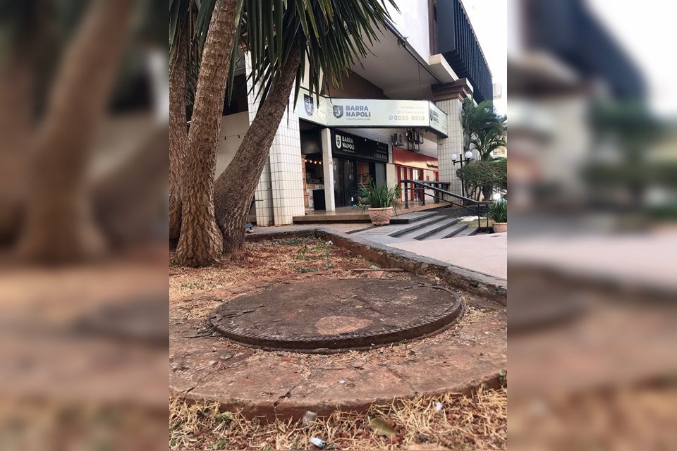 Tampa de bueiro, árvore atrás e comércio