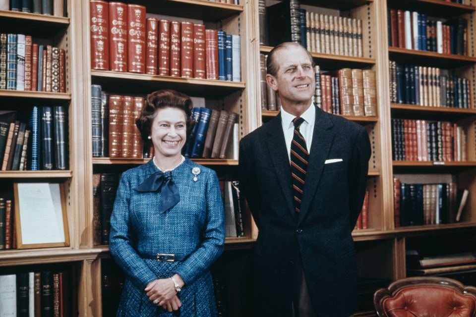 Foto colorida. Mulher de vestido azul ao lado de homem de terno. Ao fundo, prateleiras com livros