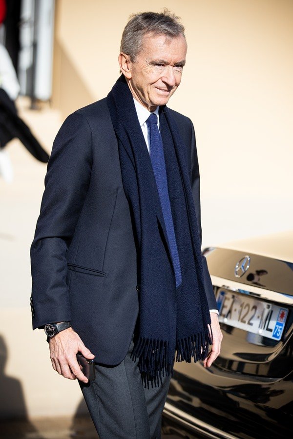 O empresário francês Bernard Arnault, um homem idoso e branco, de cabelo liso, branco e curto, posando para foto na saída do desfile da marca Dior, da qual é dono. Ele usa camisa branca de botão, um blazer escuro e gravata. A foto é de 2020.