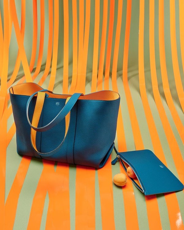 Foto de campanha da marca Moynat, que vende artigos em couro. Na imagem é possível ver uma bolsa estilo sacola de compras, larga, e uma carteira. Ambas são de couro azul e possuem a parte interna laranja