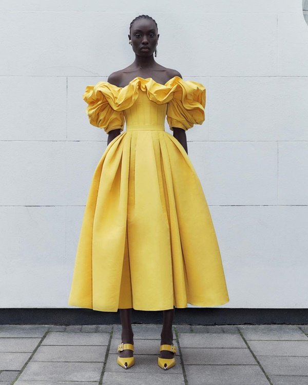 Mulher negra e jovem, de cabelo trançado, posando para foto em um fundo branco. Ela usa um vestido amarelo estilo princesa da marca de luxo Alexander McQueen.