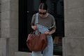Mulher branca e jovem, de cabelo castanho liso amarrado em um coque, mexendo na própria bolsa enquanto anda na rua. A bolsa é grande, estilo sacola, feita de couro marrom. A mulher usa uma camiseta cinza e um macacão jeans, além de óculos escuros pretos.