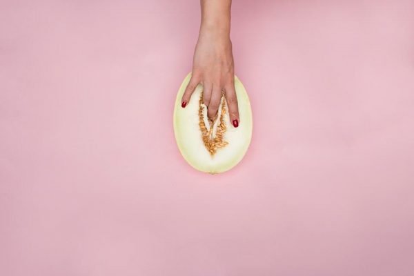 Foto colorida de uma mão feminina com as unhas vermelhas com os dedos dentro de uma fatia de melão em um fundo cor-de-rosa - Metrópoles