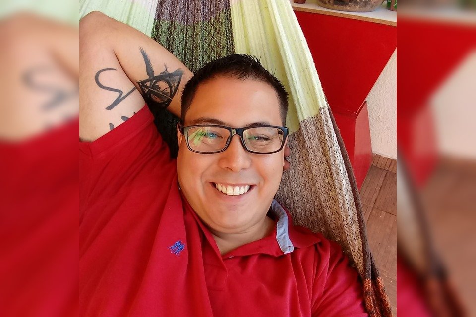 Homem com camisa vermelha, óculos e tatuagem no braço deitado em rede e sorrindo