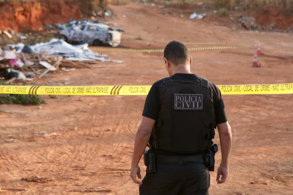 Policial fardado de preto, de costas, em frente a fita amarela em terreno de barro