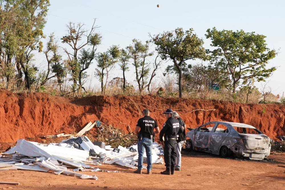 Policiais em pé em terreno de barro com carro queimado