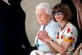 A Rainha Elizabeth II sentada ao lado de Anna Wintour, jornalista de moda da revista Vogue, durante a Semana de Moda de Londres em 2018. A monarca usa um casaco azul claro de tweed e a jornalista está com um vestido branco com flores vermelhas e amarelas e um óculos escuro.