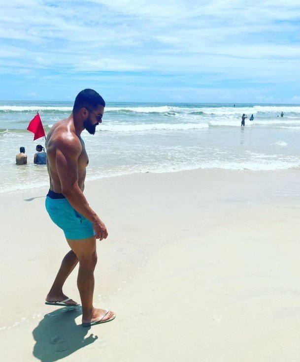 Homem de cabelos negros e curtos e barba longa caminha descalço em areia da praia. Veste short azul. Mar ao fundo. Foto colorida
