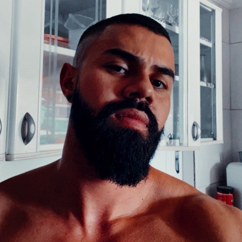 Homem de cabelos negros e curtos e barba longa olha para dentro. Está sem camisa e tem um armário atrás