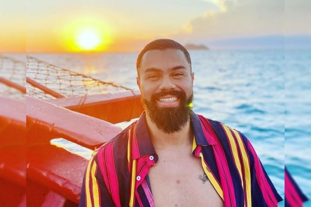 Homem de cabelos negros e curtos sorri para foto em praia. Veste camisa colorida aberta