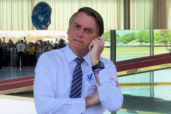 O presidente Bolsonaro fala enquanto coça a cabeça com o braço cruzado, na entrada do Palácio do Planalto. No reflexo do espelho atrás de si, é possível ver uma multidão de apoaidores de verde amarelo em sua frente - Metrópoles