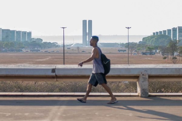 Homem de mochila preta, regata cinza, bermuda cinza-escuro e chinelo caminha em calçada na região central de Brasília, com Congresso Nacional ao fundo