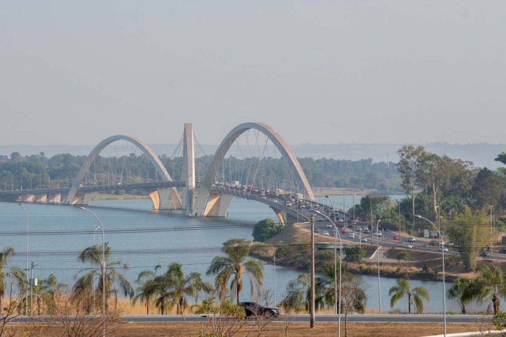 Ponte JK, sobre o Lago Paranoá, em Brasília, com céu marcado por névoa seca; carros trafegam pela ponte e por via em primeiro plano; grama seca e palmeiras em primeiro plano
