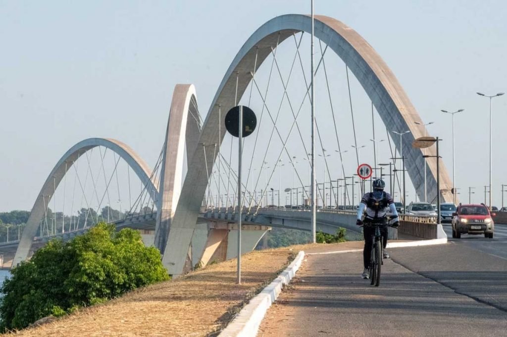 Ciclista pedala, com capacete e casaco azul e preto, com Ponte JK ao fundo. Há um arbusto à esquerda, veículos passando pela via, uma placa de trânsito ao fundo, além de postes ao longo da ponte