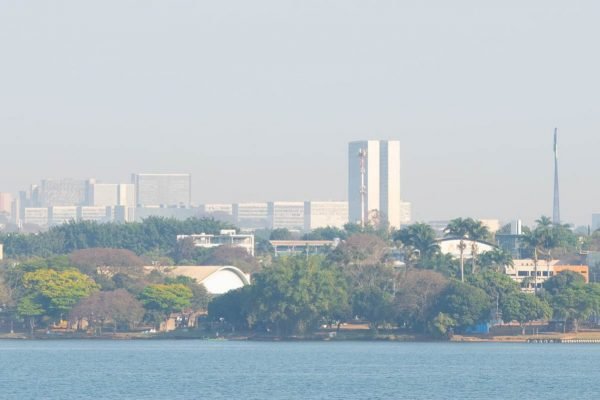 Névoa seca em Brasília, com prédios da região central da cidade, como Congresso Nacional e ministérios, ao fundo e Lago Paranoá em primeiro plano; árvores e grama seca aparecem à frente dos edifícios