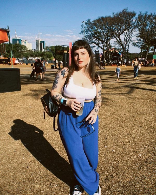 Mulher branca e jovem, com tatuagens no braço, posando para foto no festival Coma, em Brasília, na região do Eixo Monumental. Ela usa um top branco esportivo, calça azul folgada e uma mochila preta de couro