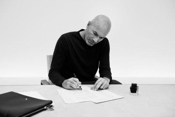 Campanha de divulgação da marca de luxo Montblanc. A foto, feita em preto e branco, mostra o jogador de futebol Zinédine Zidane, um homem branco, de meia idade e careca, com objetos da marca alemã. Ele mostra itens como canetas, pastas e cadernos.