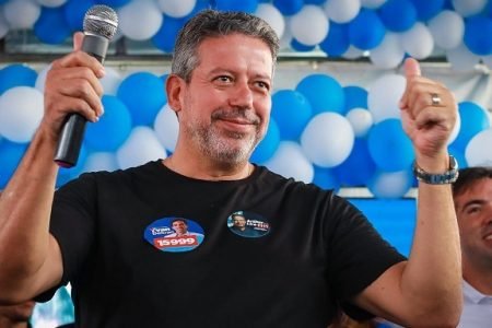 Arthur Lira, presidente da Câmara dos Deputados, sorri em cima de palanque durante campanha. Ele segura microfone na mão esquerda e atras é possívle ver balões azuis e brancos - Metrópoles