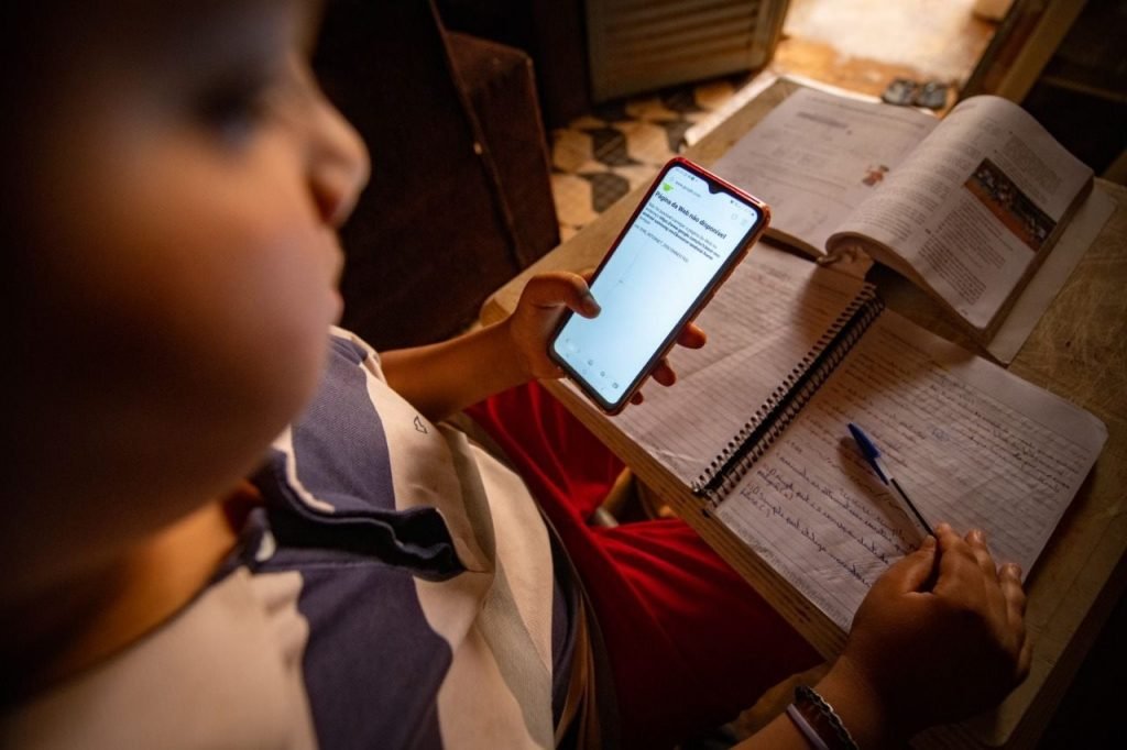 Menino com bochechas grandes sentado, olhando para baixo onde há um caderno, uma caneta e um celular sem internet na mesa