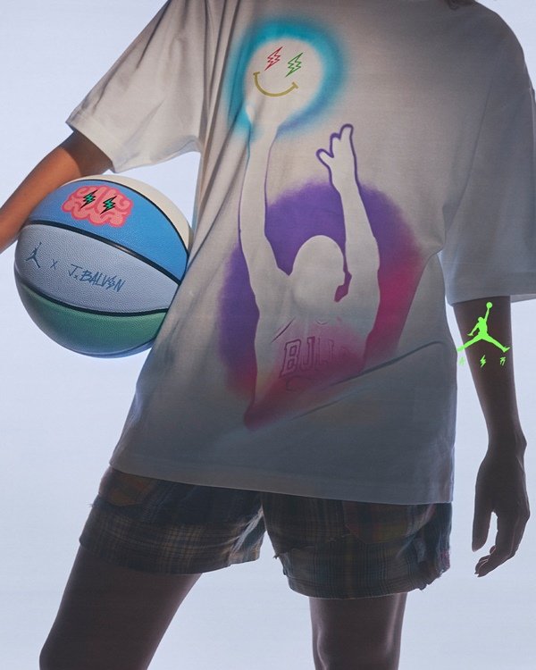 Campanha de divulgação da parceria entre a marca Jordan, do jogador de basquete Michael Jordan, com o cantor J Balvin. Os itens são feitos pela Nike. Na foto, é possível ver uma pessoa de camiseta branca e shorts lilás segurando uma bola de basquete com as listras pintadas de branco, azul e verde claro.