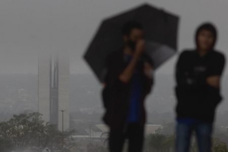 homens com guarda chuva caminham proximo a torre de tv com vista para o Congresso nacional