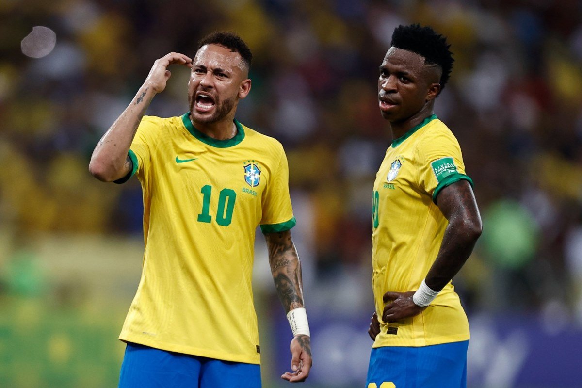 Foto de Neymar ao lado de Vini Jr. durante partida para o Brasil, os dois estão usando a camisa amarela da Seleção e Neymar reclama com os braço perto do rosto, enquanto Vini Jr. oberserva a situação