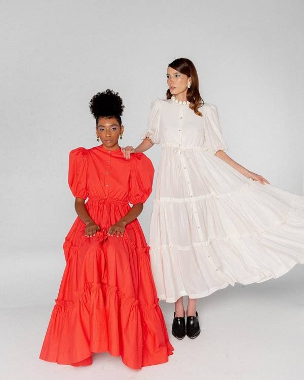 Modelos usam vestidos longos com mangas bufantes. Um é laranja e o outro é branco, da marca Lucila Pena