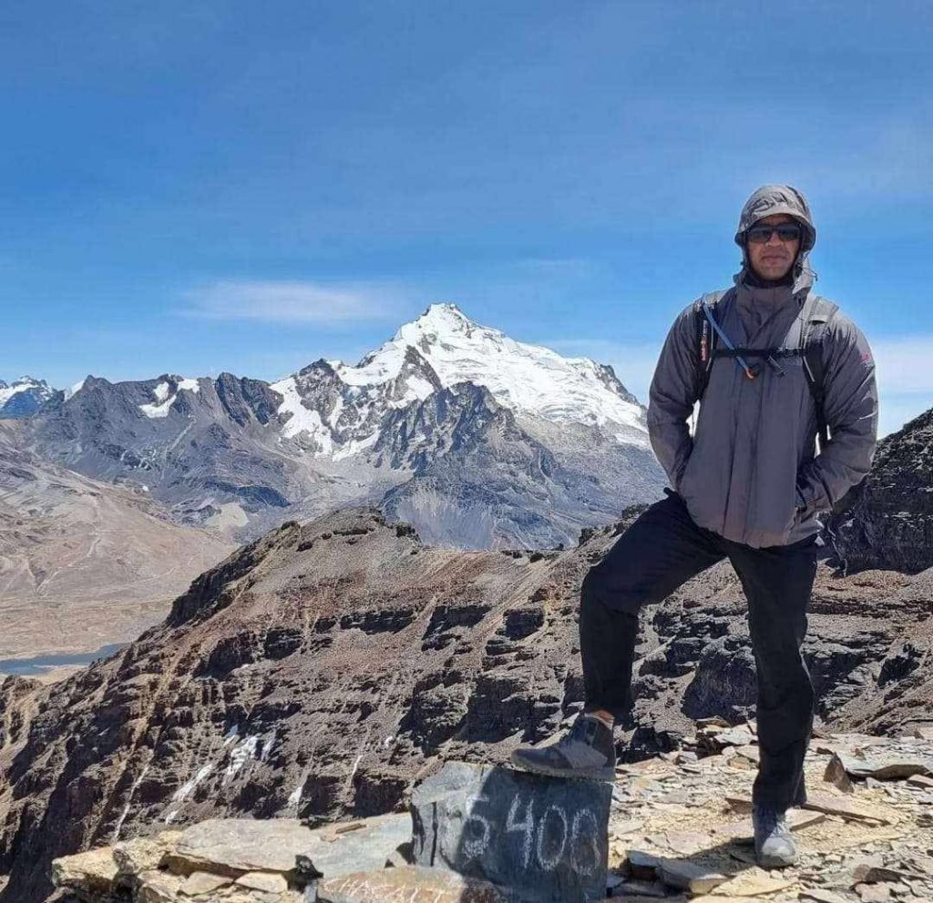 Foto colorida. Homem de casaco cinza posa para foto em montanha