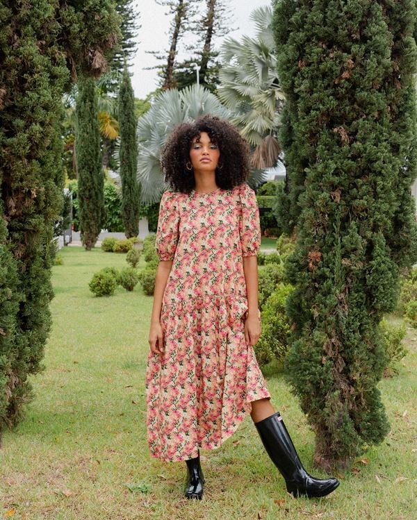 Mulher negra e jovem, de cabelo cacheado, posando para foto em um jardim. Ela usa um vestido longo de estampa florida rosa e uma bota longa preta estilo galocha 