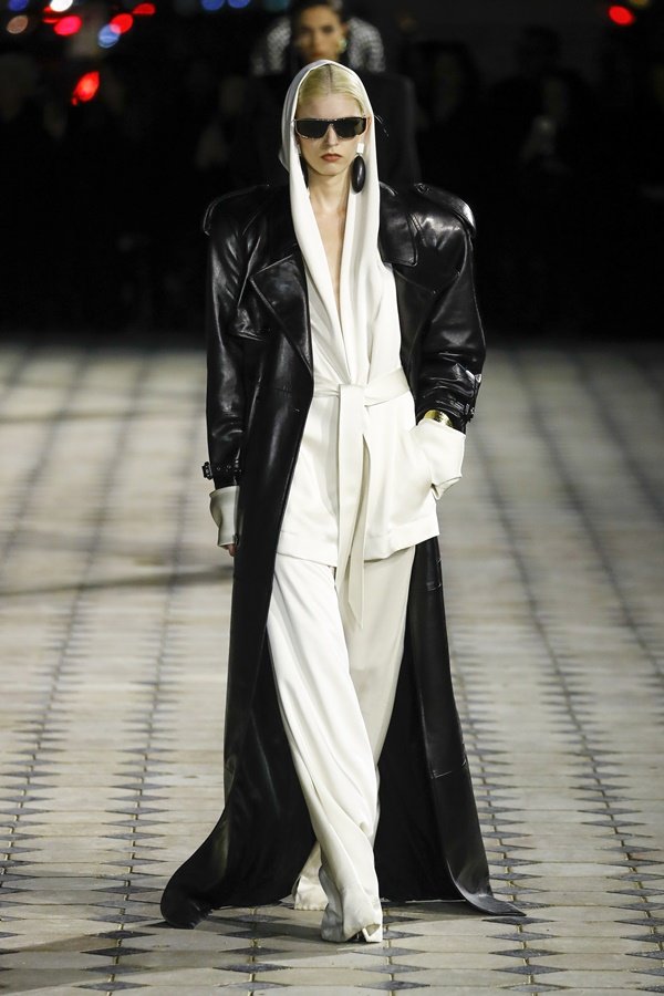 Uma modelo caminha na passarela durante o desfile de moda da Saint Laurent, coleção de Primavera/Verão 2023, como parte da Paris Fashion Week em 28 de setembro de 2022. A modelo é uma mulher branca de cabelo liso amarrado e usa um conjunto de calça e blazer brancos e, por cima, um casaco estilo sobretudo de couro preto