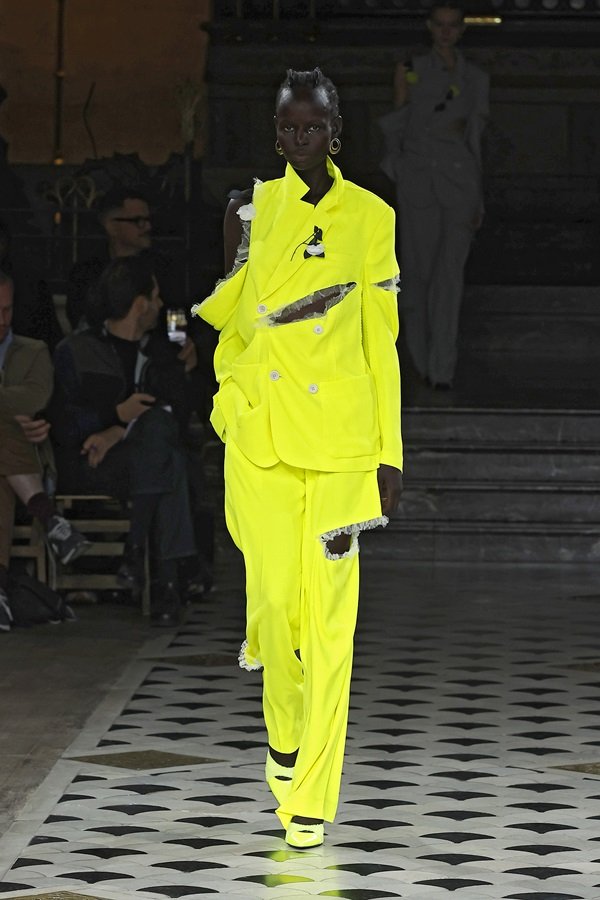 Uma modelo caminha na passarela durante o desfile de moda da Undercover, coleção de Primavera/Verão 2023, como parte da Paris Fashion Week em setembro de 2022. A modelo é uma mulher negra e jovem, de cabelo raspado, e usa um conjunto de blazer a calça em amarelo neon.