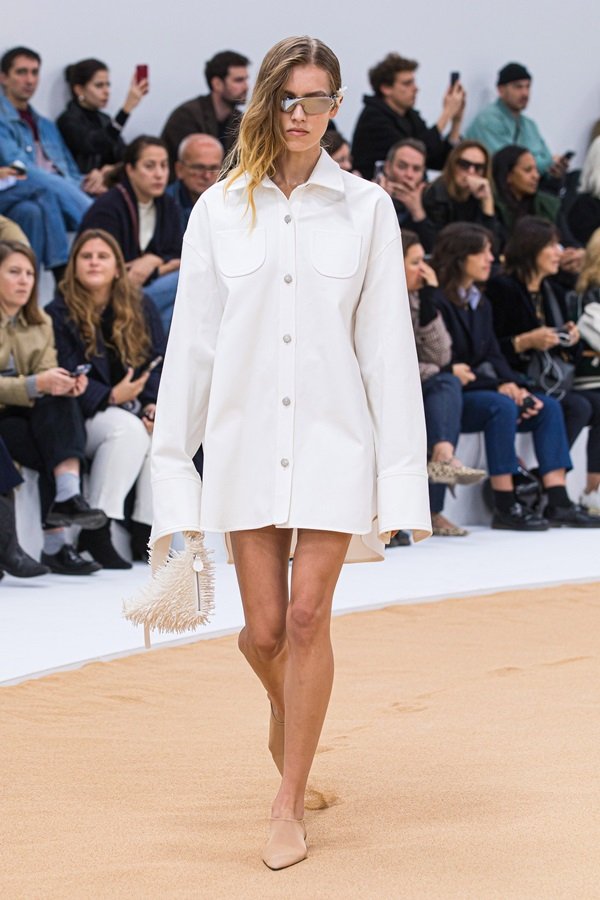 Uma modelo caminha na passarela durante o desfile de moda da Courrèges, coleção de Primavera/Verão 2023, como parte da Paris Fashion Week em setembro de 2022. A modelo é uma mulher branca e jovem, de cabelo longo loiro, e usa uma camisa branca de botão como vestido e óculos escuros espelhados.
