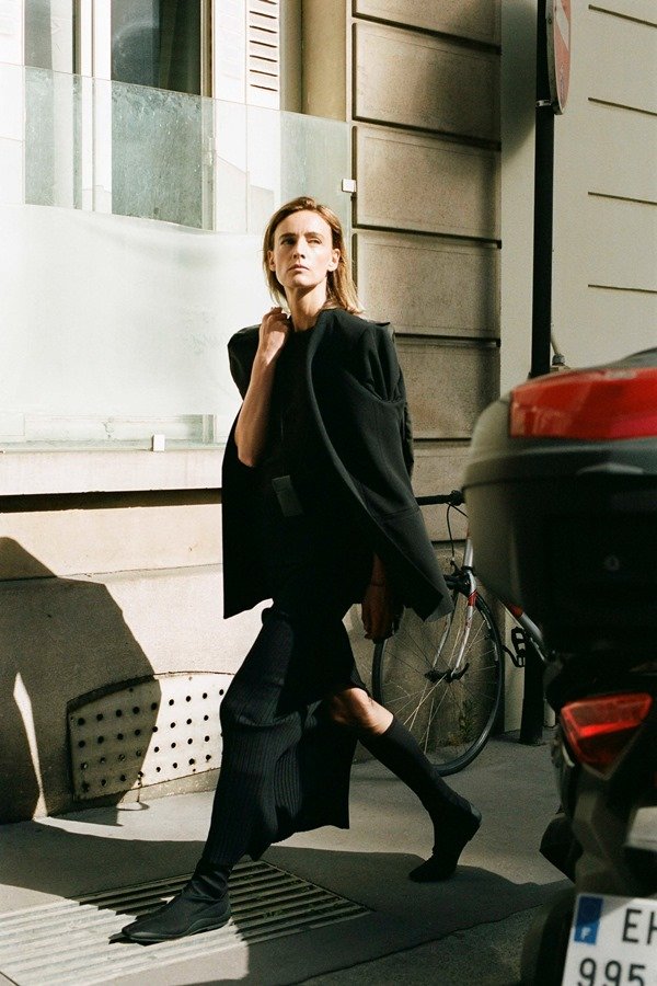 Uma modelo caminha na passarela durante o desfile de moda da The Row, coleção de Primavera/Verão 2023, como parte da Paris Fashion Week em setembro de 2022. A modelo é uma mulher branca e jovem, de cabelo longo loiro, e usa uma camisa preta de botão, blazer e calça de alfaiataria pretos.