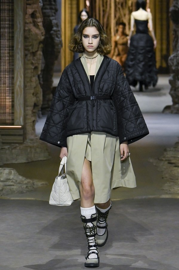Uma modelo caminha na passarela durante o desfile de moda da Dior, coleção de Primavera/Verão 2023, como parte da Paris Fashion Week em setembro de 2022. A modelo é uma mulher branca e jovem, de cabelo castanho liso e curto, e usa um vestido bege e um casaco acolchoado preto.